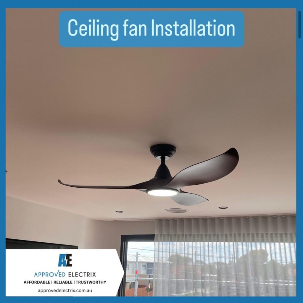 A modern black ceiling fan with three curved blades and an integrated light is installed in a contemporary room. The ceiling is light-colored, and the room features large windows with sheer curtains, allowing natural light to enter. The image is framed with a blue border, featuring the Approved Electrix logo and the text “Ceiling fan Installation.”